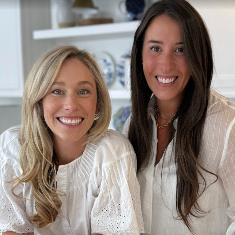Two women smiling in a bright kitchen, both wearing white short-sleeve blouse with eyelet lace and gathered neckline. One has blonde hair and the other has long brown hair. They are the founders of Baby Bump